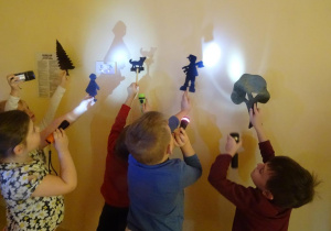 Pięcioro dzieci trzyma w ręku sylwety ludzi, zwierząt i roślin na które święcą latarkami tworząc cień na ścianie.